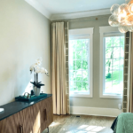 A modern bedroom with light gray walls, large windows with beige curtains, a wooden sideboard, a potted orchid, and a contemporary chandelier with spherical lights.